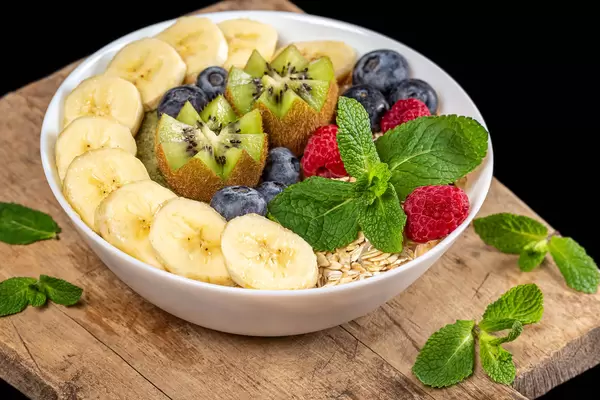 Bowl of oatmeal with blueberries, banana, kiwi, raspberries and mint on wooden kitchen board, close-up