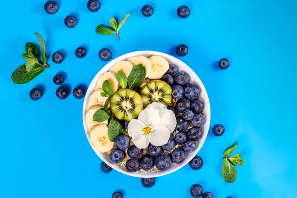 Bowl of oatmeal with blueberry, banana, kiwi edible flower blossom on blue background with fresh mint leaves, top view
