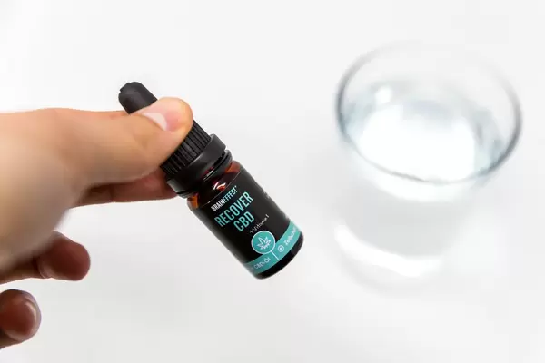 Braineffet Recover CBD Öl + Vitamin E Fläschchen vor Wasserglas aus weißem Untergrund