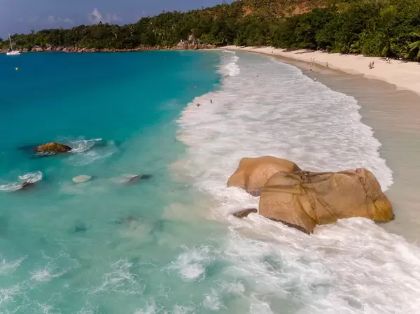 Brauner Granitstein umgeben von den Wellen des Indischen Ozeans an der Nordküste Anse Lazio mit Touristen im Hintergrund auf Praslin, Seychellen