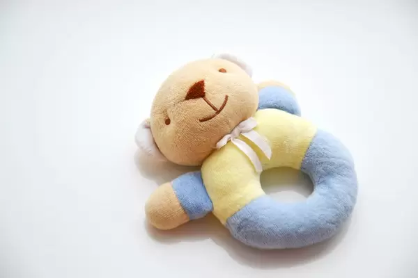 Brauner Teddybär mit weißer Schleife um den Hals und blau-gelbem Körper in Form eines Beißrings vor weißem Hintergrund