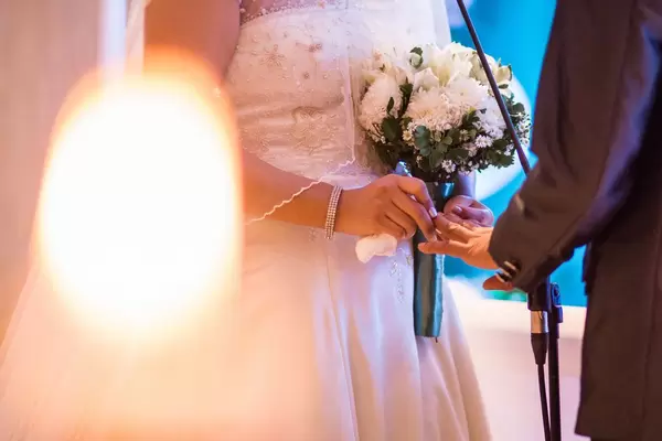 Braut in weißem Kleid hält Brautstrauß und steckt zukünftigem Ehemann den Ehering an