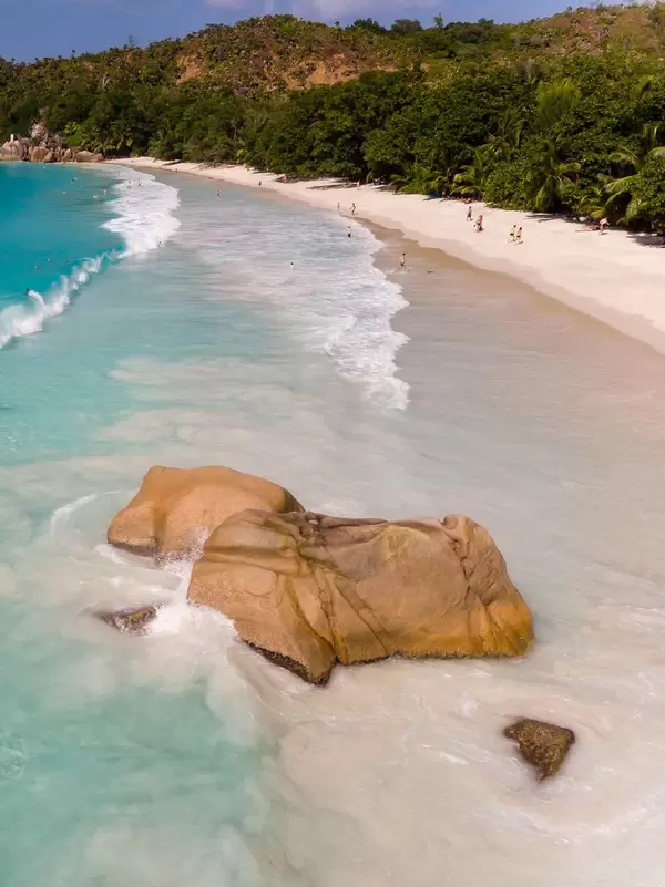 Brown granite rock covered by waves of the Indian Ocean with tourists on the white beach Anse Lazio in the background in Praslin, Seychelles