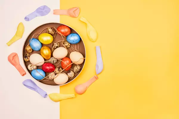 Bunte Ostereier und Wachteleier auf dem Teller, Aufnahme von oben mit Luftballons und gelbem Hintergrund