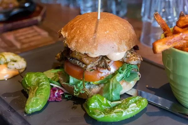 Burger on a wooden skewer, with tomato, salat, meat patty and sauce, next to green pepperonis and sweet potato chips in the restaurant