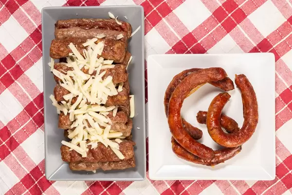 Cevapcici bestreut mit Käsestücken in Schale neben Teller mit saftigen Würstchen auf rot-weißem Tuch