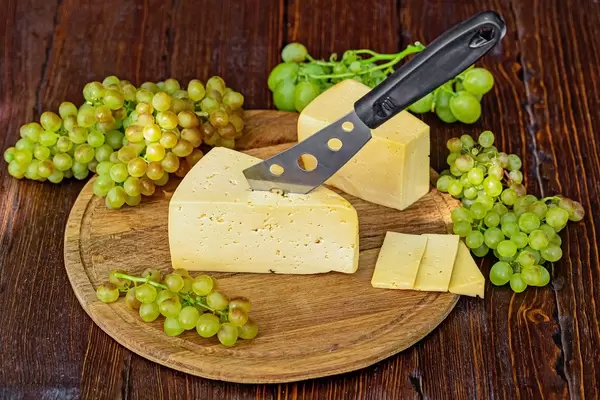 Cheese knife stuck in a piece of cheese on a round wooden board with ripe fresh grapes