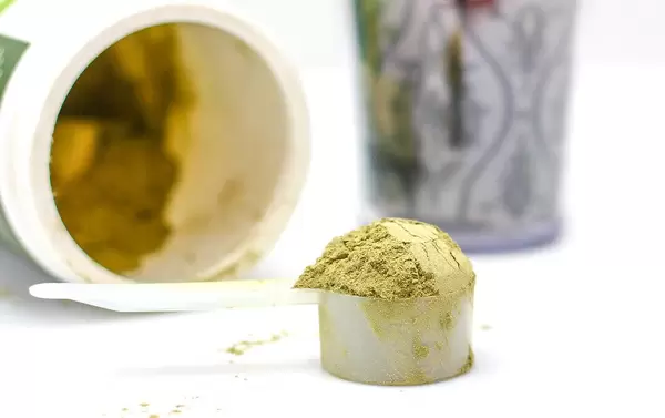Close Up Bokeh Photo of Green Protein Powder in Plastic Measure Spoon with Container and Glass in the Background