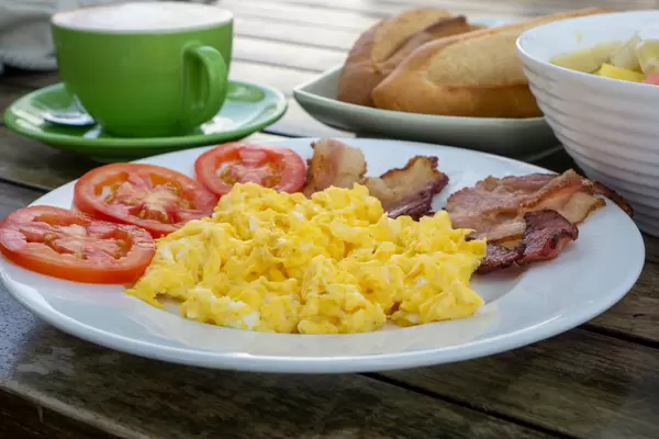 Close Up Food Photo of Breakfast with Scrambled Eggs, Bacon Strips, Tomatoes, Baguette, Cappuccino and Fruit Bowl on a Wooden Table