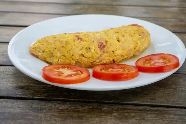 Close Up Food Photo of Egg Omelette with Onions and Bacon on a White Plate with Tomato Slices on a Wooden Table