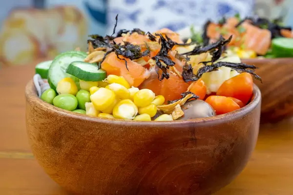 Close Up Food Photo of Hawaiian Poke Bowl with Corn, Tomatoes, Cucumber, Salmon, Fried Onions and Dried Seeweed in a Wooden Bowl