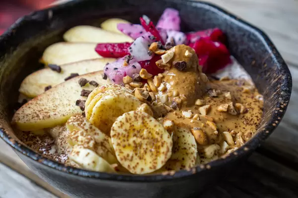 Close Up Food Photo of Healthy Breakfast Bowl with Oatmeal, Cinnamon, Apple, Banana, Dragon Fruit, Chocolate Cubes, Peanuts and Peanut Butter in a Black Ceramic Bowl
