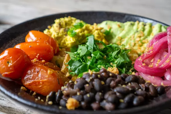 Close Up Food Photo of Healthy Meal with Grilled Cherry Tomatoes, Beans, Scrambled Tofu with Spring Onions, Avocado and Baked Potatoes on a Black Ceramic Plate