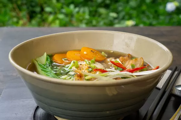 Close Up Food Photo of Japanese Ramen Noodle Soup with Pork, Spring Onions, Bok Choy and Sliced Carrots in a Ceramic Bowl on a Wooden Table with Chopsticks
