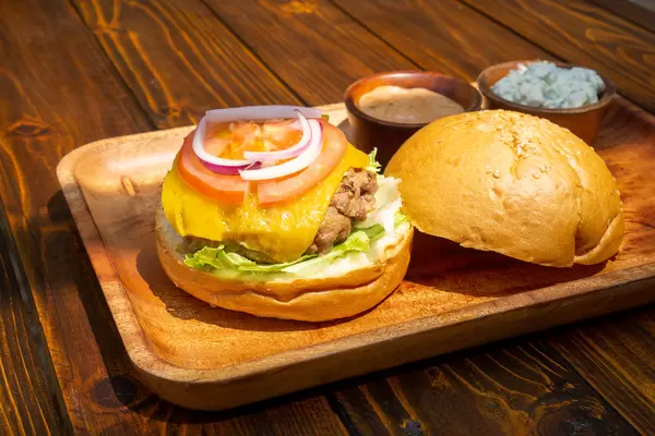 Close Up Food Photo of Kofta Burger with Lamb Meat Patty, Lettuce, Melted Cheese, Tomato, Onion and Sauces on Side on a Wooden Plate in a Restaurant