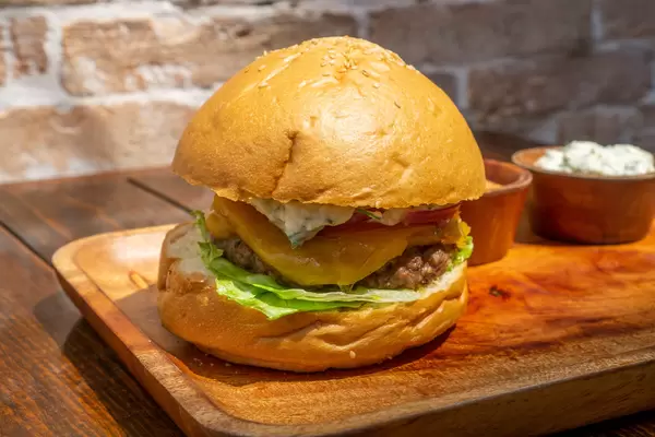Close Up Food Photo of Kofta Burger with Sesame Buns, Tzatziki and Cocktail Sauce, Lamb Meat Patty, Lettuce, Raw Onions and Melted Cheese on a Wooden Plate with Brick Wall in the Background