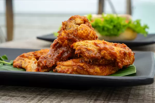 Close Up Food Photo of Marinated Spicy Chicken Wings on a Black Plate with a Sandwich in the Background on a Wooden Table