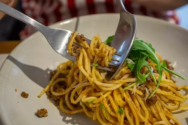 Close Up Food Photo of Person using Fork and Spoon to eat Spaghetti Bolognese with Minced Meat, Tomato, Grated Cheese and Arugula in a Restaurant