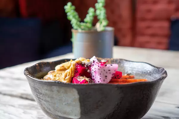 Close Up Food Photo of Red and White Dragon Fruit Cubes in a Healthy Breakfast Bowl with Oatmeal, Peanut Butter, Nuts and Apple with a Table Plant in the Background