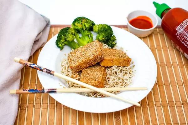Close Up Food Photo of Sesame Tofu, Broccoli, Ramen Noodles and Sriracha Sauce on Bamboo Tray with Chopsticks
