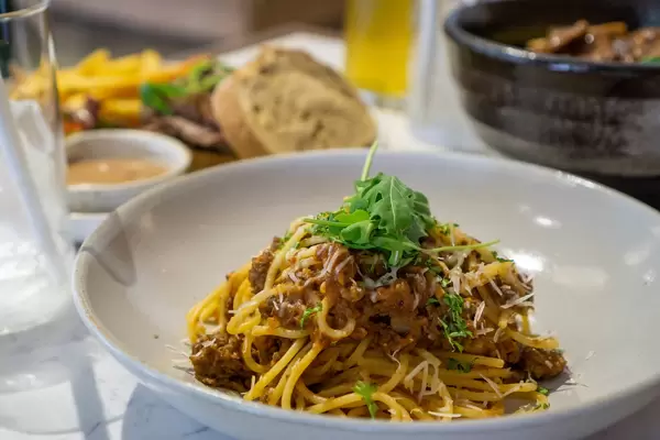 Close Up Food Photo of Spaghetti Bolognese with Tomato and Minced Meat topped with Grated Cheese and Arugula with other Dishes in the Background