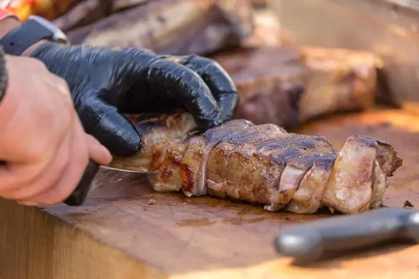 Close-up of a man cutting a large piece of OFYR barbecue meat into pieces on a wooden table