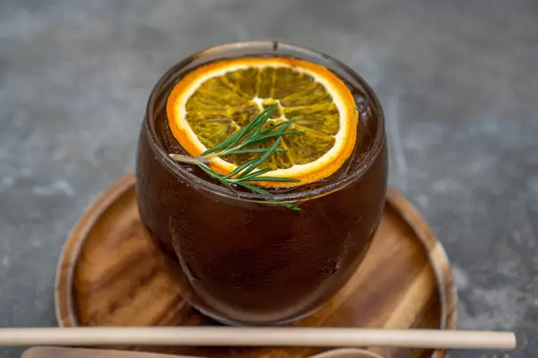 Close Up of Burnt Orange Cold Brew Coffee with Slice of Orange and Rosemary on a Wooden Plate with Paper Straw and Wooden Spoon