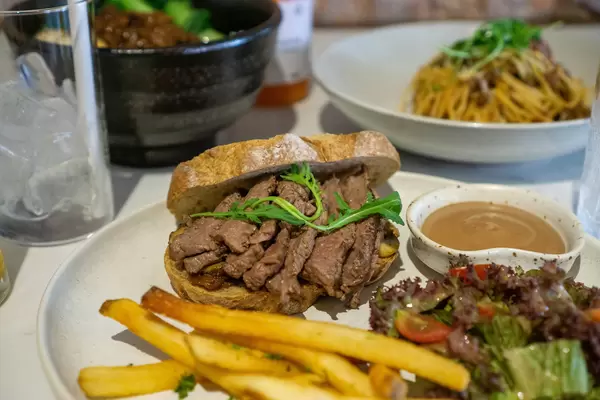 Close Up Photo of Food Plate with Steak Slices on Bread Slice with Caramelized Onions, French Fries and Salad as Side Dish and other Dishes in the Background