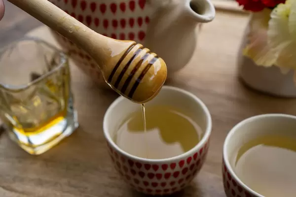 Close Up Photo of Honey dripping from a Honey Spoon into a Small Ceramic Cup with Green Tea on a Wooden Tray