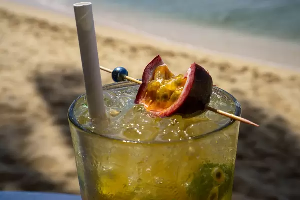 Close Up Photo of Mojito with Passion Fruit Seeds, Fresh Passion Fruit, Ice Cubes, Mint, Lime, Sugar and Paper Straw in a Cocktail Glass with Beach in the Background