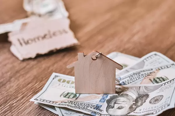 Close-up shot of wooden house toy on a stack of US dollars and homeless note at the background