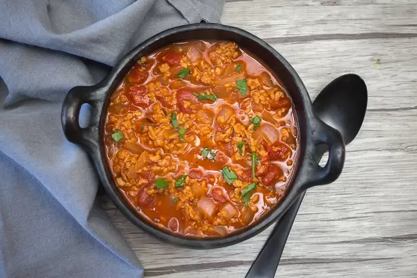 Close Up Top View Food Photo of Ground Beef Chili in Black Ceramic Clay Pot next to Spoon and Kitchen Cloth on Wooden Table