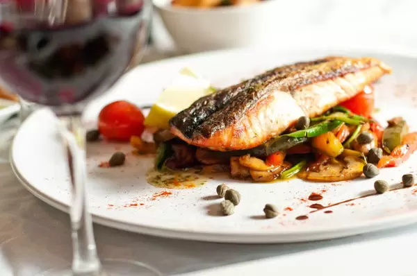 Closeup photograph of a plate with fish and vegetables with an unsharp glass of wine in the foreground.