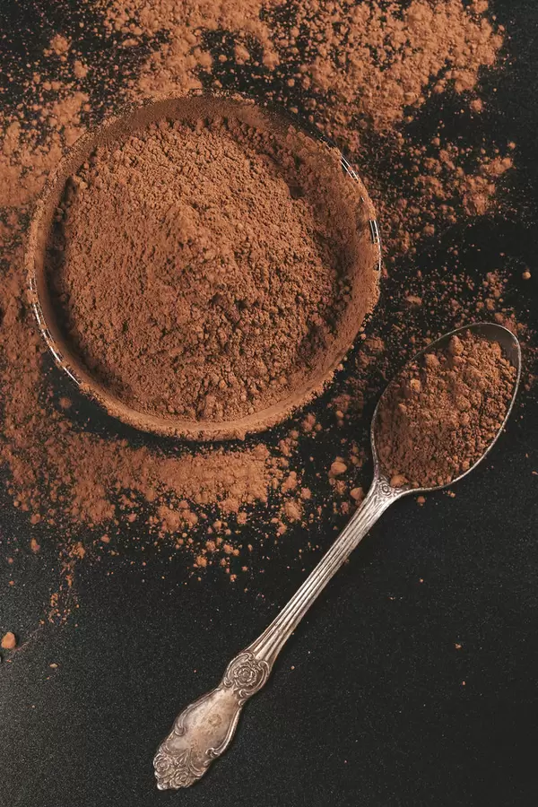 Cocoa powder in bowl and spoon on dark background, top view