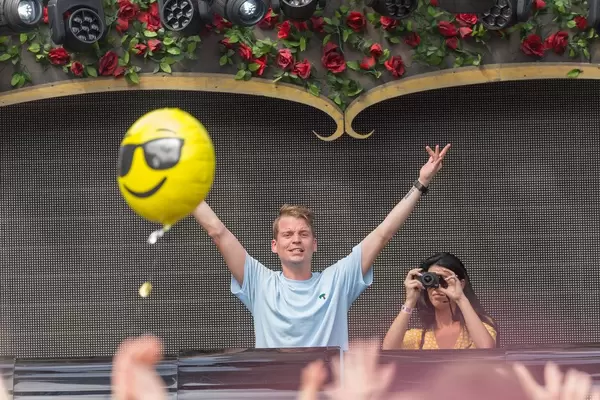 Conro dances with lifted hands while someone flies a smiley balloon and a woman photographs the party from the DJ stage