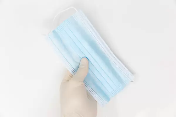 Corona, zweite Welle: Hand mit Einweghandschuh hält eine blaue OP-Maske vor weißem Hintergrund