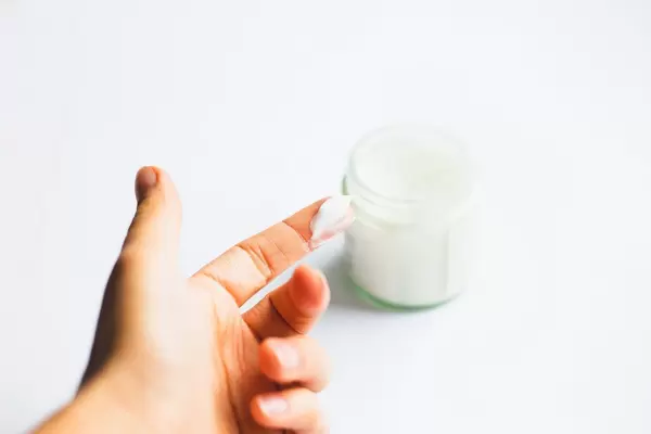 Creme auf dem Finger einer Frau mit einem Glas Creme im Hintergrund - Nahaufnahme