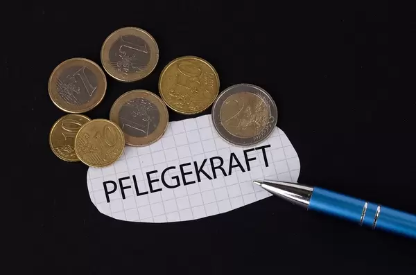 Das Konzept von der Pflegekraft: Pflegekraft Text auf einem Blatt Papier mit einigen Münzen und einem blauen Stift im schwarzen Hintergrund