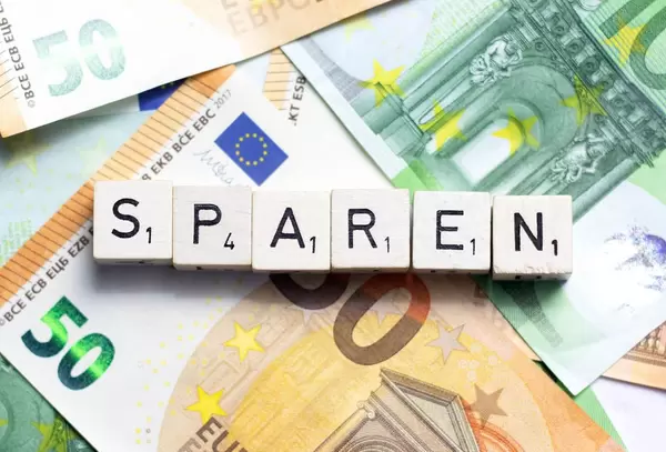 Das Wort Sparen auf Würfelbuchstaben mit Euroscheinen im Hintergrund