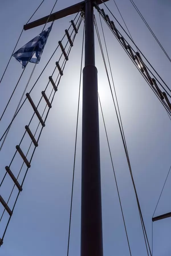 Detail of a sailboat with Greek flag and the sun hidden behind the mast, seen from below