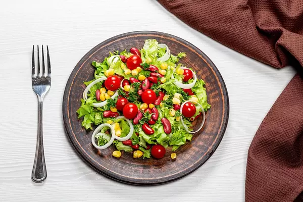 Diät-Salat mit buntem Gemüse und roten Bohnen auf einem braunen Teller, mit einer Gabel und Küchentuch, auf dem Tisch