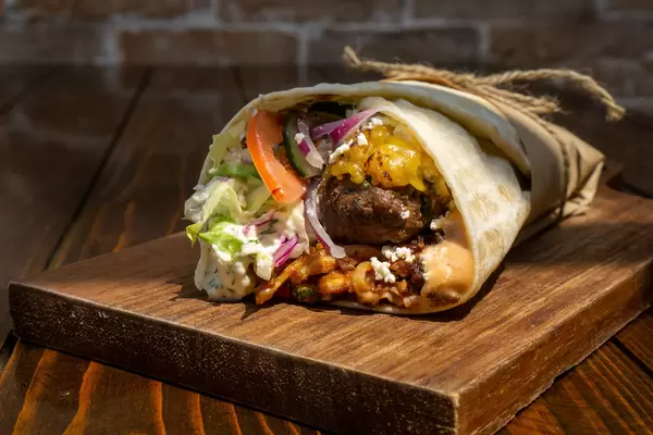 Döner Dürum Wrap als Fladenbrot gefüllt mit Lammfleisch, Käse, Feta, Zwiebeln, Tomaten, Gurken, hausgemachtes Tzatziki, Cocktailsoße, Salat und Zucchini auf einem Holzbrett in einem Imbiss Nahaufnahme