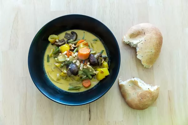 Draufsicht von gelbem Curry mit Ofengemüse und einem Brötchen