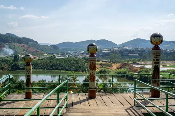 Drei Figuren in verschiedenen Größen auf einer Holz-Aussichtsplattform mit Dörfern, Bergen und einem See im Hintergrund in Da Lat, Vietnam