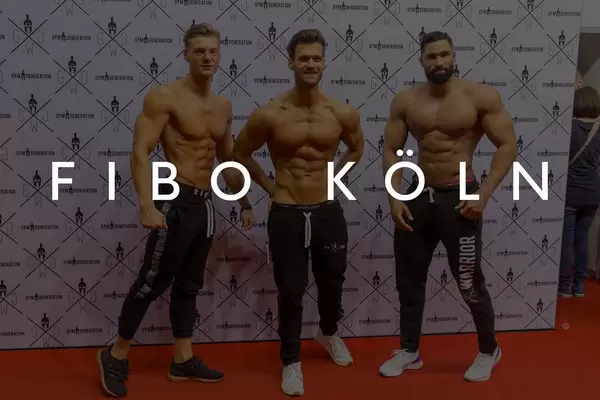 Drei sportliche Männer mit nacktem Oberkörper zeigen ihre Muskeln und Sixpacks auf dem roten Teppich, mit dem Bildtitel "Fibo Köln"