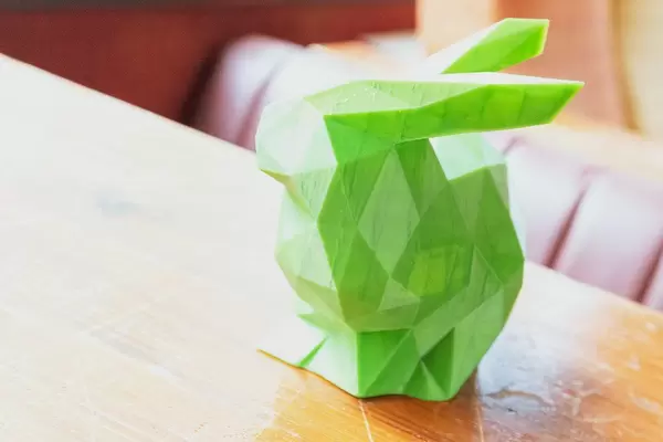 Dreidimensionaler ausgedruckter grüner Hase aus dem 3D-Drucker, steht im Sonnenlicht auf einem Holztisch