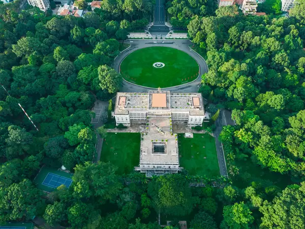 Drohnenaufnahme vom Unabhängigkeitspalast mit Helikopter umgeben von einem Park mit vielen Bäumen im Stadtzentrum von Ho Chi Minh Stadt, Vietnam