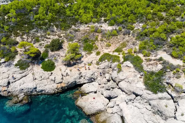 Drohnenaufnahme zeigt Touristen auf einer Gesteinsküste, beim Erkunden der Sehenswürdigkeit Bekiris Höhle, auf der saronischen Insel Spetses