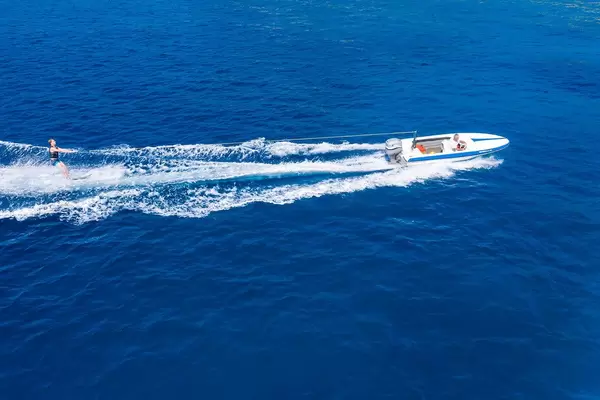 Drone picture of a water sports enthusiast on water skis, pulled by a motor sport boat across the blue sea and breaking white waves