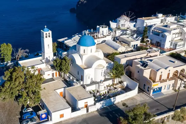 Drone shot. White church and bell tower with typical blue domes, cemetery and windmill in Santorini